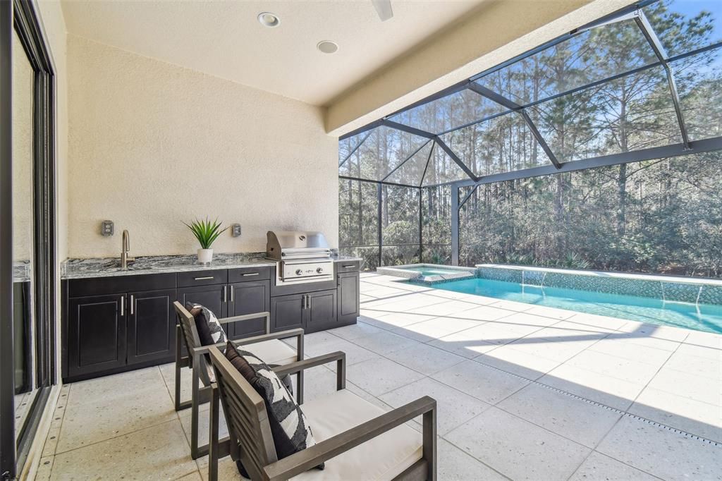 The outdoor kitchen has a sink, grill, stone counters and wood cabinets.