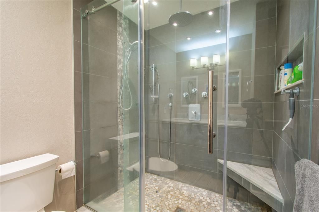 dual sink mirrored vanity, a walk-in barn door glass shower featuring body sprays, a rainfall shower head, and a bench for a spa-like experience.