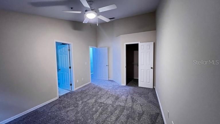 The primary bedroom room with a neutral color palette, is freshly carpeted in a soft gray. A modern ceiling fan ensures airflow and comfort. The room features multiple doors, including one leading to the primary bathroom with natural light filtering through the visible open door and a large walk-in closet and wooden window shade.