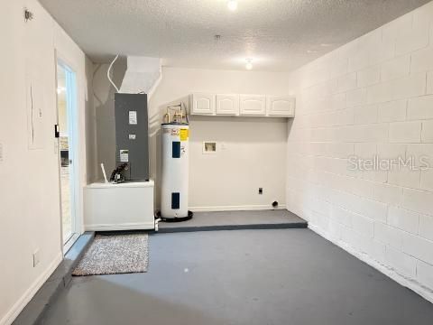 The laundry area is a practical utility area within the garage, featuring a clean, white block wall backdrop. A central HVAC unit is installed alongside a water heater, while overhead white cabinetry offers additional storage solutions. The space includes a laundry area with hookups ready, evidenced by the nearby outlets and plumbing. A neutral-toned floor mat lies in front of the door, leading into the home, indicating a transitional space from the garage interior. This functional zone is designed for efficiency, catering to household maintenance needs.