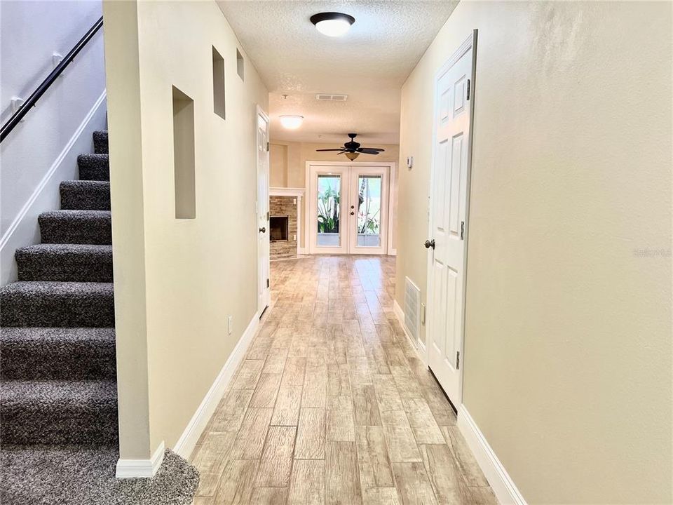 Once in the foyer, a bright and spacious hallway leads to an open living area. On the left, a staircase with new dark gray carpeting ascends to an upper level, its black handrail creating a striking contrast against the neutral wall color. The hallway features wood-look tile flooring, which extends into the inviting living space ahead, where a ceiling fan suggests the additional comfort of the living room, kitchen and dining room. Natural light appears to flow in from the front door, with sidelights illuminating the space. White doors line the right side of the hall, leading to the garage, and laundry area, and walk-in closet on the left.