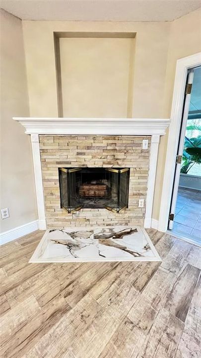 The open living space  features an elegant, modern fireplace as its centerpiece. The fireplace has a clean, classic white mantel surrounding a stone tile facade in neutral tones, which complements the wood-look flooring. Above the fireplace is a recessed niche, potentially for art or a television. The hearth is uniquely designed with what appears to be marble tiles, adding a touch of luxury. The natural light from an adjacent room and the reflective glass doors of the fireplace amplify the room's brightness. The space exudes a contemporary yet homey feel, with ample room for furnishings and decor to enhance the inviting atmosphere.
