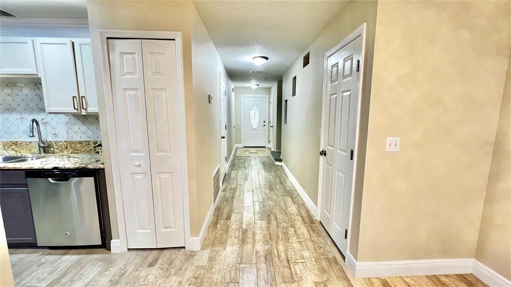 This hallway exudes a welcoming feel with its light wood-look flooring that contrasts pleasantly with the neutral wall tones, creating a bright and open feel. To one side, the hallway grants a glimpse of the kitchen, featuring white upper cabinetry, dark lower cabinets, and a stylish backsplash that complements the granite countertops. Overhead, recessed lighting ensures the space is well-lit, while the view down the hall leads to a beautifully detailed front door, promising a warm welcome for visitors. The corridor is lined with multiple doors, suggesting ample storage or room entrances, making the space both functional and inviting.