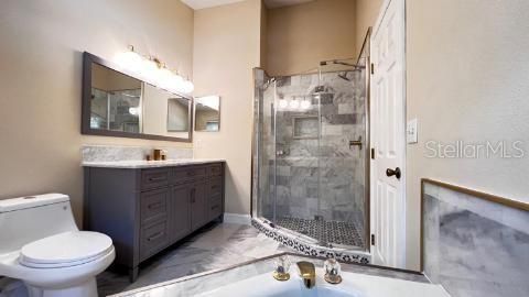Enjoy this tastefully designed bathroom featuring a spacious glass-enclosed shower with marble-like tiles that provide a luxurious feel. A contemporary gray vanity with ample cabinetry is topped with a contrasting light countertop, complemented by a large mirror with modern lighting above. The gold-tone fixtures add a touch of elegance. The room is completed with a matching tiled floor, enhancing the cohesive and upscale appearance.