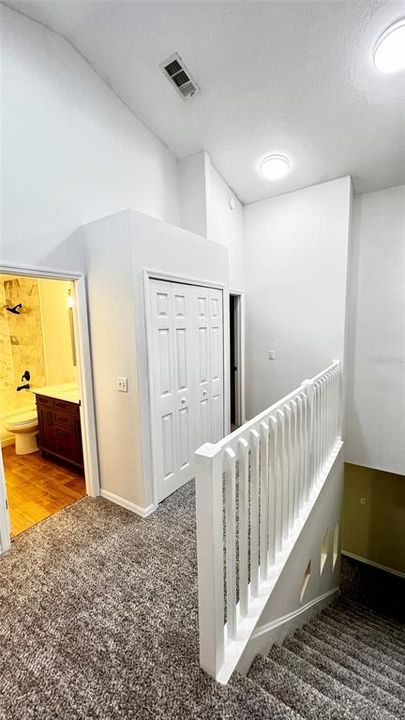 The upper hallway features a well-lit, high-ceiling space at the top of a staircase, characterized by clean, white finishes. On the left, an open door leads to a bathroom with a wooden vanity and a mirror, showing a glimpse of a shower and a toilet. To the right, the space is open to the staircase, bordered by a white balustrade that gives a sense of openness while maintaining safety. The carpeted flooring suggests comfort and warmth in this residential setting.
