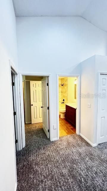 The upper landing that leads to the primary, second and third bedrooms features a well-lit, high-vaulted ceiling space at the top of a staircase, characterized by clean, white finishes. On the left, an open door leads to a bathroom with a wooden vanity and a mirror, showing a glimpse of a shower and toilet. To the right, the space is open to the staircase, bordered by a white balustrade that gives a sense of openness while maintaining safety. The carpeted flooring suggests comfort and warmth in this residential setting.