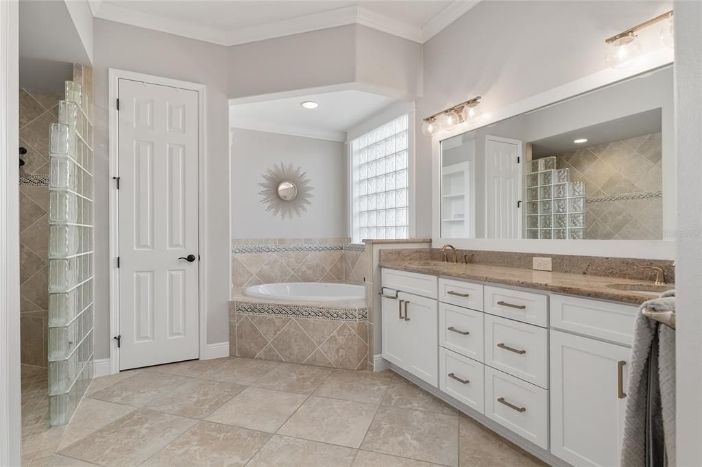 ENSUITE BATHROOM view shows ROMAN SHOWER w/ GLASS BLOCK WALL (L), linen closet, jetted SOAKING TUB (center), & DUAL SINKS/VANITY w/ GRANITE COUNTERTOPS (R). Note updated gold hardware on cabinets, gold faucets & gold lighting fixtures.