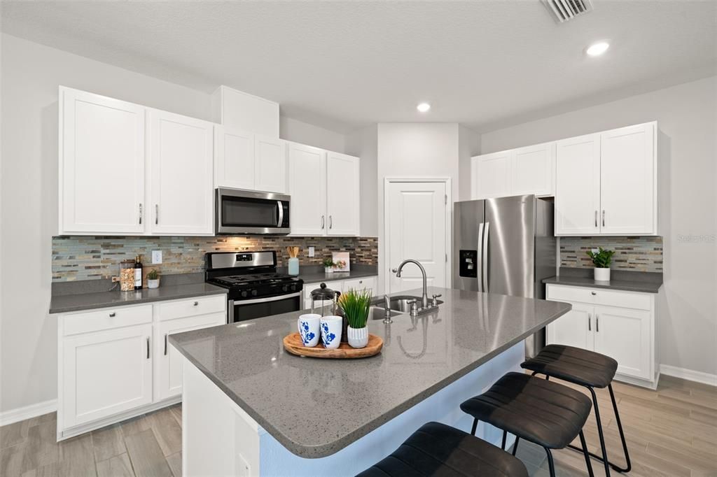 Upgraded kitchen featuring quartz countertops, walk-in pantry, gas stove, stainless steel appliances, cabinet pulls, neutral backsplash and a large island.