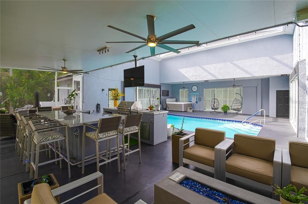 View of the expansive covered lanai with outdoor kitchen and additional seating with lighted gas burning center table. Notice the lovely wooded view