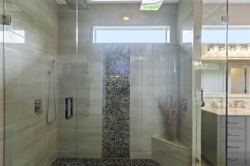 View of updated walk in shower with 12" square double rainfall shower heads, body jets, frameless custom glass enclosure, pebble mosaic floor and wall time and built in seat.