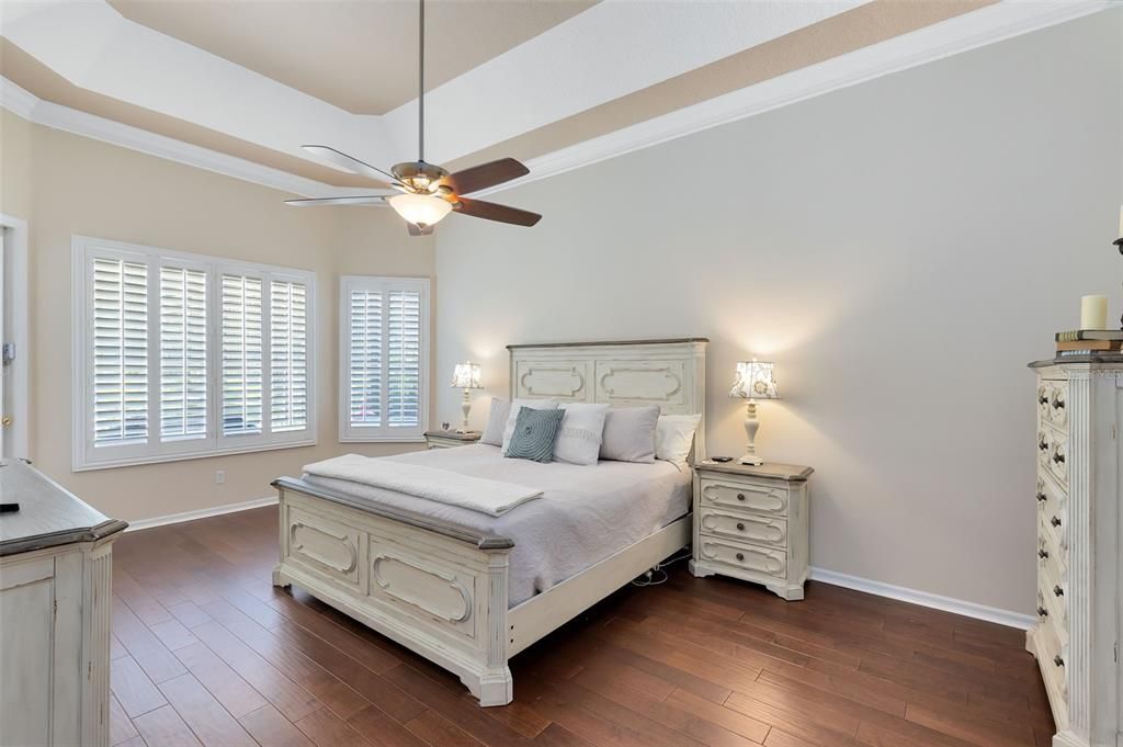 PRIMARY BEDROOM WITH TRAY CEILING, WOOD FLOORING AND PLANTATION SHUTTERS WITH VIEW OF THE LANAI AND POOL