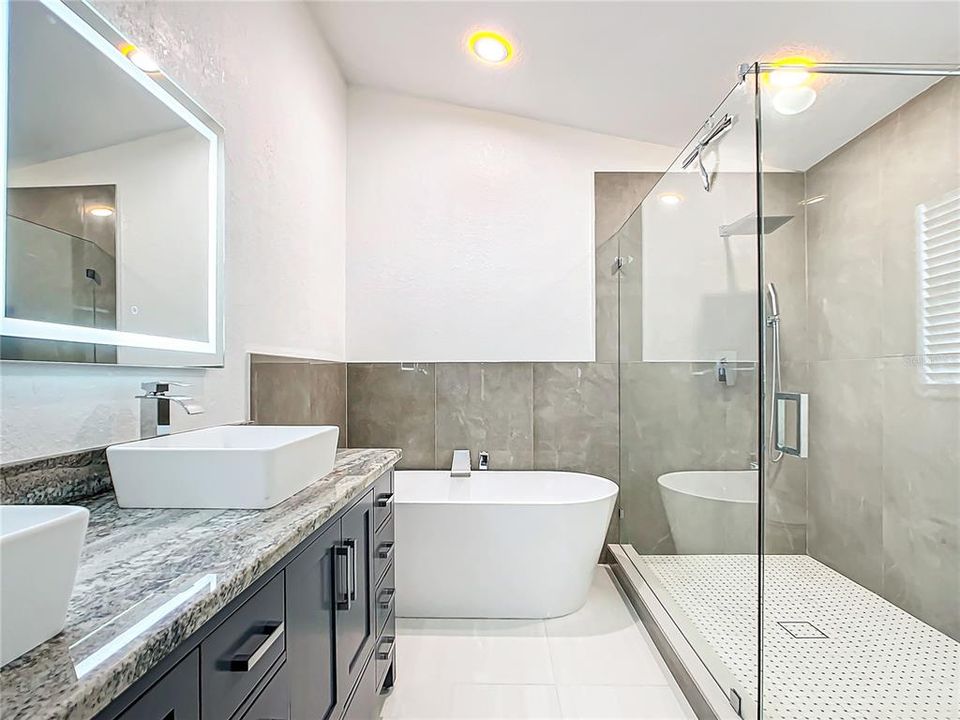 The En-suite primary bathroom has been beautifully remodeled with a soaking tub, separate shower, updated dual-vessel sinks, granite countertops, and illuminated defogging mirrors.