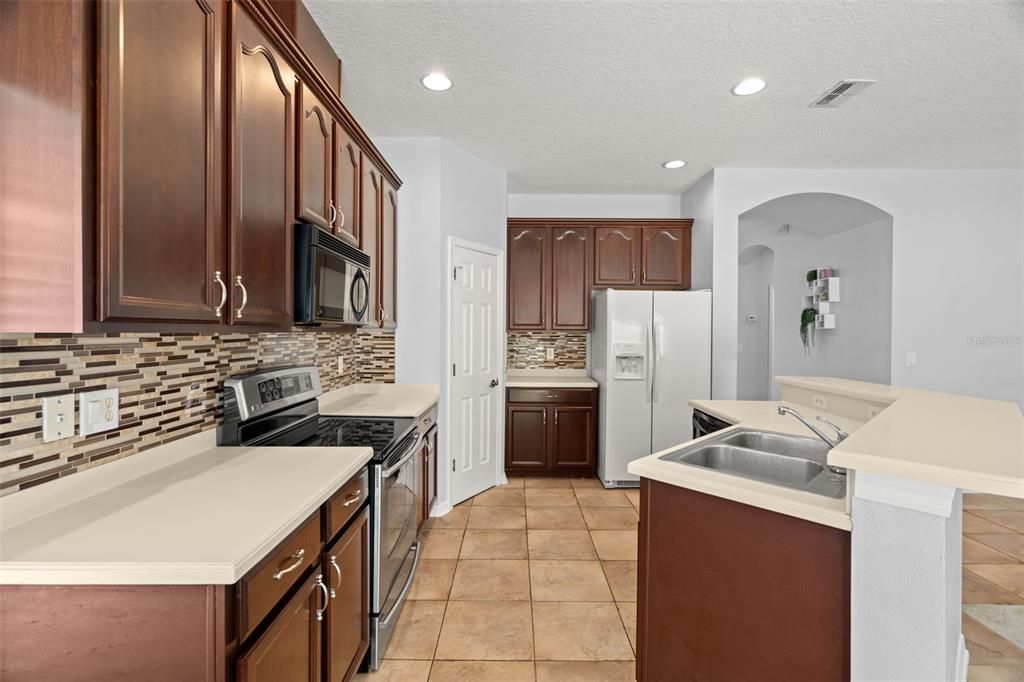The kitchen features solid surface countertops, glass tile backsplash, ample and tall cabinets, pantry closet, kitchen island with single basin sink and dishwasher, brand new smooth cooktop range and white double door refrigerator.