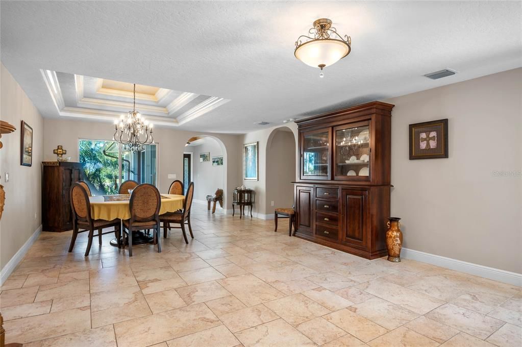 The elegant dining area at 6215 S Tropical Trl, Merritt Island, FL, is a perfect blend of sophistication and comfort. This spacious room features a beautiful tray ceiling with crown molding and a luxurious chandelier, providing an opulent atmosphere for dining and entertaining. The large sliding glass doors allow natural light to flood the space, offering picturesque views of the lush tropical surroundings. The rich wood furniture, including a stately china cabinet and dining table, adds warmth and character to the room. The travertine tile flooring continues seamlessly, enhancing the overall aesthetic and flow of the home.