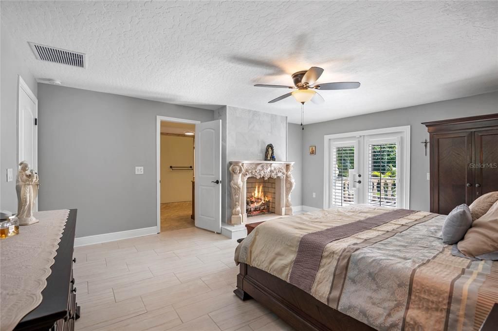 The master bedroom continues to impress with its blend of comfort and elegance. The room features a cozy fireplace with an ornate mantel, adding warmth and charm. French doors with plantation shutters lead to the outdoor patio, allowing natural light to flood the room and providing easy access to outdoor living. The ceiling fan and recessed lighting offer both functionality and style. The spacious layout includes access to a walk-in closet through an open door, enhancing the room's practicality and luxury. The neutral color palette and tile flooring create a serene and inviting atmosphere.