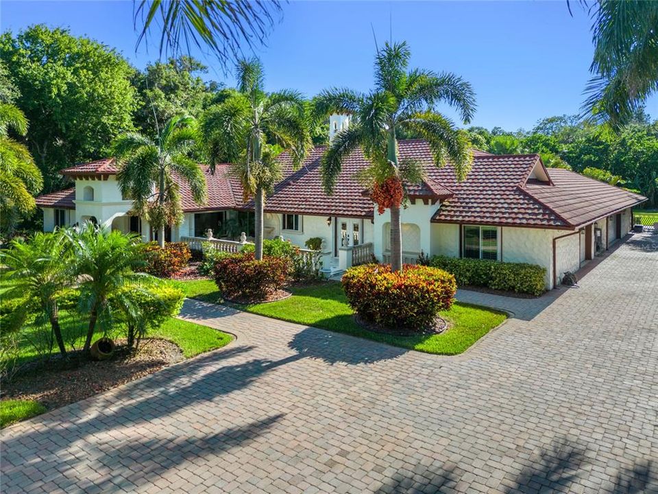 This angle showcases the expansive driveway leading to a spacious three-car garage, perfectly blending functionality with style. The home's exterior features a pristine stucco finish complemented by a red tile roof, creating a harmonious and inviting aesthetic. Lush landscaping, including meticulously trimmed bushes and towering palm trees, adds a touch of natural elegance to the property. The clear blue sky overhead highlights the vibrant and serene environment, making this residence a true Floridian paradise.