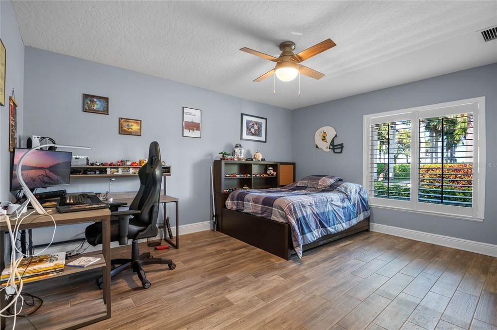 This cozy bedroom features wood-look tile flooring, large windows with shutters for natural light and privacy, and a ceiling fan for added comfort.
