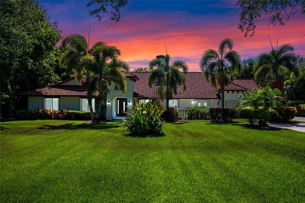 This stunning property is showcased against a breathtaking sunset, highlighting its elegant facade and lush landscaping. The expansive front lawn, adorned with vibrant greenery and tropical palm trees, leads to the grand entrance. The Mediterranean-style architecture, complete with a tile roof and arched doorway, exudes sophistication and charm, making this home a true tropical oasis.