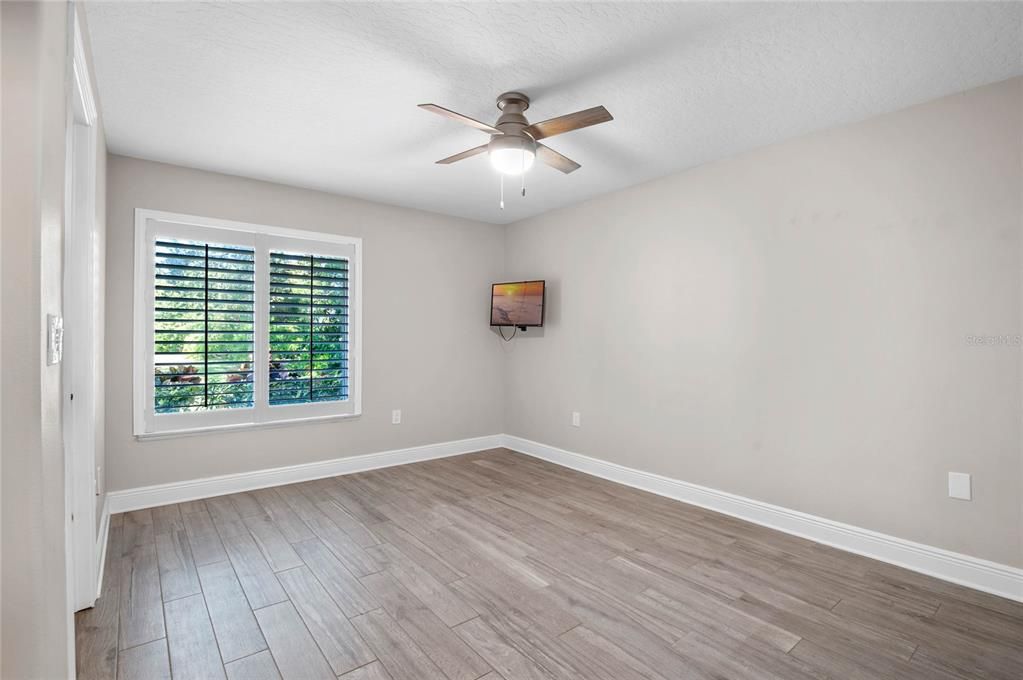 This bright and airy bedroom features a clean, neutral color palette that enhances the natural light streaming through the window. The room is equipped with plantation shutters for privacy and light control. The ceiling fan with a light fixture adds comfort and functionality, while the wood-look tile flooring provides a stylish and easy-to-maintain surface. This versatile space can be used as a bedroom, home office, or guest room, offering both comfort and flexibility.