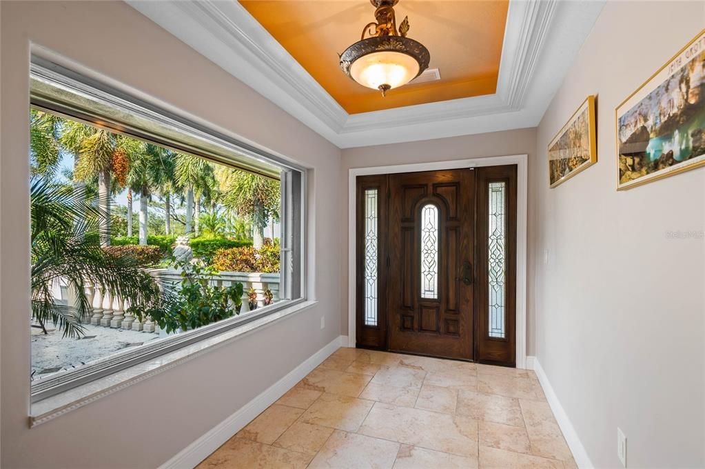 The foyer greets you with a beautiful blend of natural light and sophisticated design. The large picture window offers a stunning view of the lush tropical landscaping and majestic palm trees, creating a seamless connection between the indoors and outdoors. The intricate glass inserts on the wooden front door enhance the space with their artistic detail. The travertine tile flooring and tray ceiling with a stylish light fixture add a touch of luxury to this welcoming entryway, setting the tone for the exquisite interiors that await.