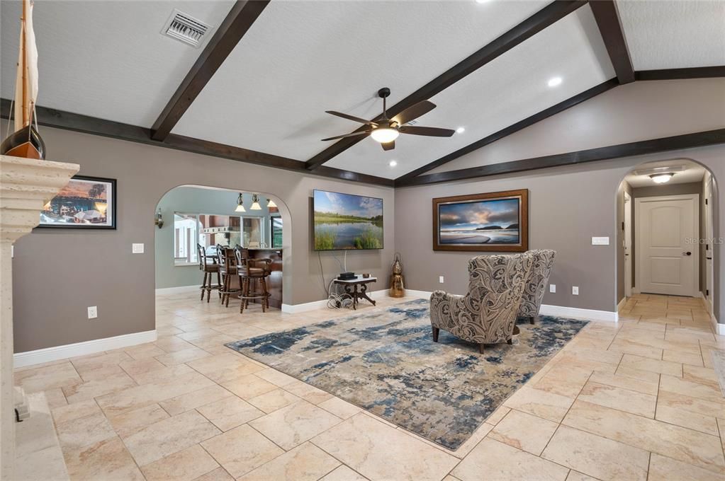 Step into the spacious living room featuring stunning architectural details such as exposed wooden beams on the ceiling, adding a touch of rustic elegance. The room is highlighted by recessed lighting and a ceiling fan, providing both style and comfort. The open floor plan allows for seamless transitions to adjacent areas, including a glimpse into the dining space through a charming arched doorway. The travertine tile flooring extends throughout, creating a cohesive and inviting ambiance. Large wall spaces offer ample room for artwork and entertainment systems, enhancing the room's functionality and aesthetic appeal.