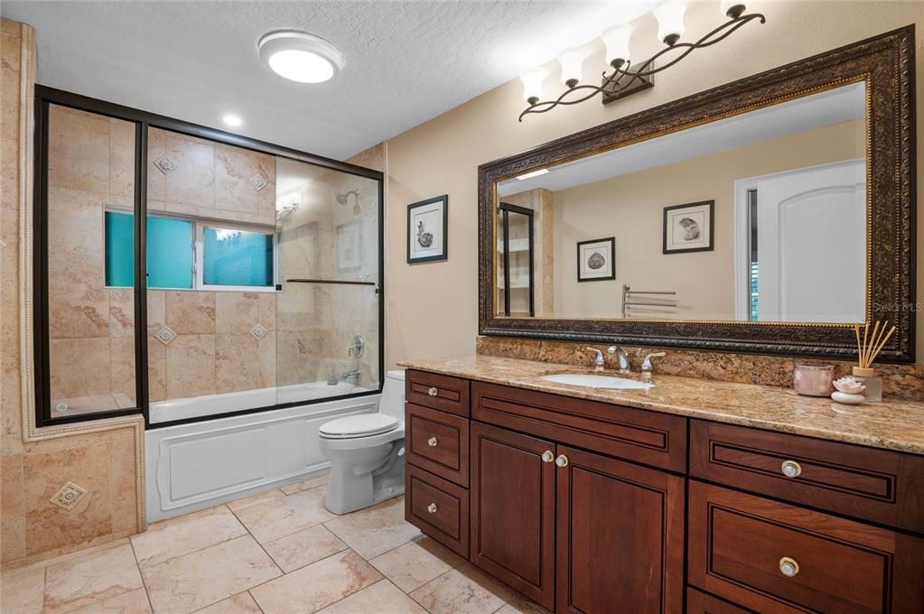 The guest bathroom is elegantly designed with high-quality finishes and fixtures. It features a large vanity with a granite countertop, providing ample storage and counter space. The framed mirror adds a touch of sophistication, complementing the warm tones of the cabinetry. The shower-tub combination is surrounded by stylish tile work, and the glass sliding doors enhance the bathroom's modern aesthetic. The room is well-lit with both recessed lighting and a decorative light fixture, creating a bright and inviting space.