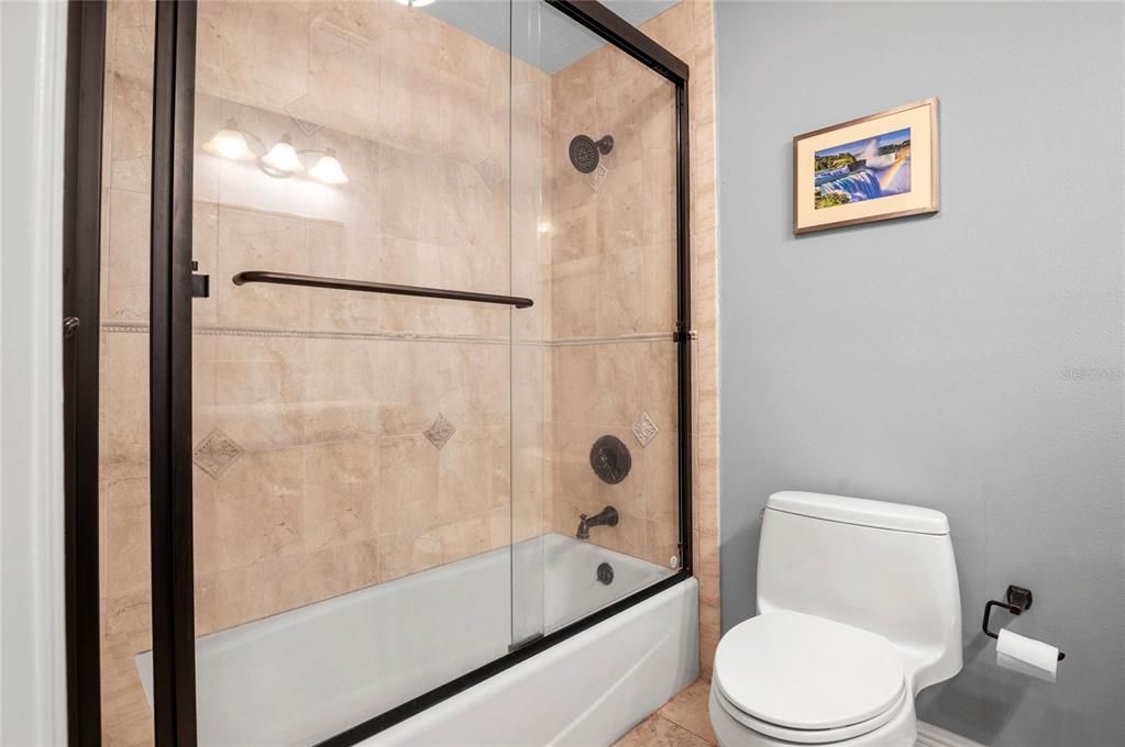 This bathroom features a combination bathtub and shower enclosed with sleek glass sliding doors. The shower walls are adorned with beige tiles that have decorative diamond accents, creating a refined look. The fixtures, including the showerhead and faucet, are finished in oil-rubbed bronze, providing a stylish contrast against the light tiles. The toilet is positioned next to the bathtub, and a framed picture adds a touch of color to the light grey walls. The overall design is modern and clean, offering both functionality and aesthetics.