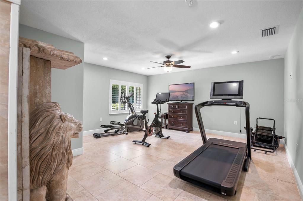 This is the in-law bedroom with separate entrance through the pool and garage.  This versatile room is currently set up as a home gym, featuring ample space, large windows with plantation shutters for natural light, and a ceiling fan for added comfort.