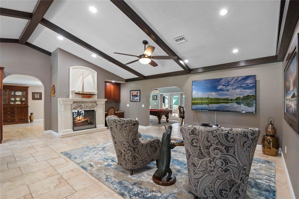 The living room seamlessly combines elegance and comfort, featuring a beautifully crafted fireplace with an ornate mantel as the centerpiece. The exposed wooden beams on the ceiling add a rustic charm, while recessed lighting and a ceiling fan ensure a bright and airy ambiance. The open layout provides easy access to adjacent areas, including a glimpse of the entertainment room through an arched doorway. The travertine tile flooring enhances the room's sophistication, making it a perfect space for both relaxation and social gatherings.