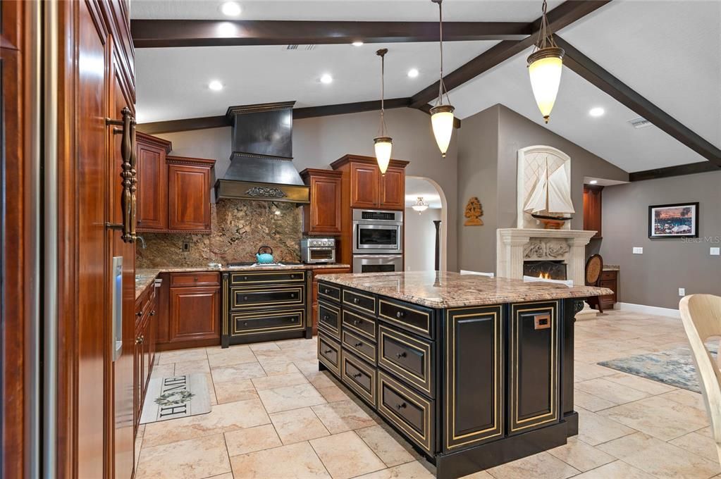 This view of the gourmet kitchen highlights the custom cabinetry and high-end appliances, including a professional-grade range with a custom hood and a built-in oven and microwave. The large center island, with its elegant granite countertop and detailed trim, offers ample storage and workspace. Pendant lighting above the island adds a stylish touch, while the exposed wooden beams on the vaulted ceiling contribute to the room's rustic charm. The travertine tile flooring ties the space together, providing a seamless and sophisticated look that complements the home's overall aesthetic.