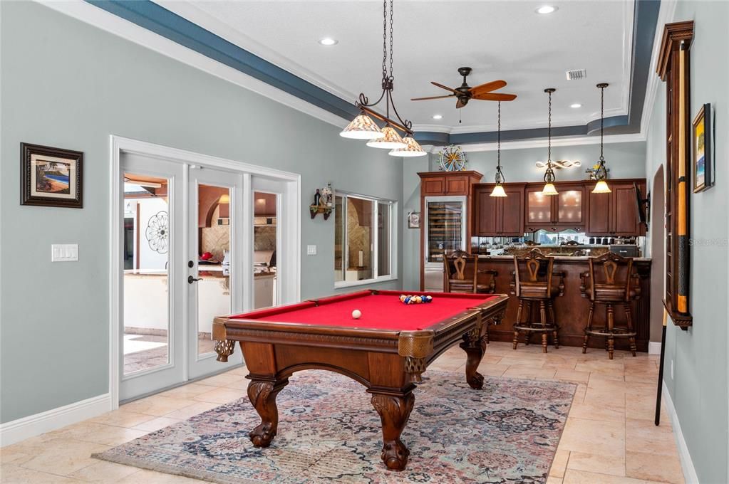 This vibrant game room offers an excellent space for recreation and relaxation, featuring a beautifully crafted pool table as the focal point. The room is adorned with a high ceiling, crown molding, and recessed lighting, creating a bright and open atmosphere. French doors lead to the outdoor patio and pool area, providing easy access to outdoor entertainment. The game room seamlessly transitions into a bar area with custom cabinetry, granite countertops, and pendant lighting, perfect for hosting guests. Large windows and a ceiling fan enhance the room's comfort and appeal, making it an ideal spot for social gatherings and leisure activities.