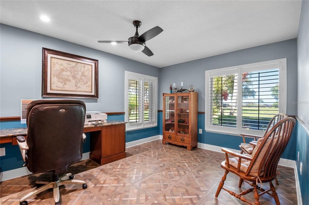 The home office offers a bright and functional workspace, featuring large windows with plantation shutters that allow natural light to fill the room. The two-tone wall color scheme, with blue on the bottom half and a neutral tone on top, adds a touch of elegance and character. The built-in desk provides ample workspace, and the ceiling fan ensures comfort while working. The room's parquet-like tile flooring adds a classic touch, making it a stylish and practical space for productivity.