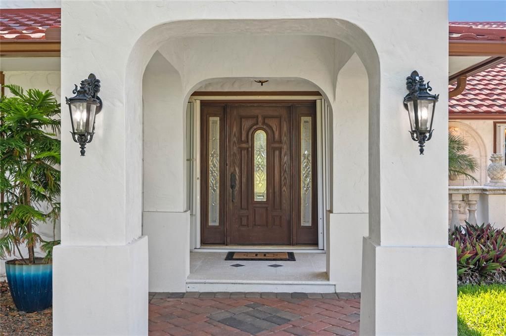 This inviting entryway features a beautifully crafted wooden front door with intricate glass inserts, adding a touch of elegance and allowing natural light to filter into the foyer. The arched stucco entry is complemented by ornate wrought-iron lanterns, providing both functionality and a classic aesthetic. The brick-paved walkway and pristine white columns create a warm and welcoming atmosphere, setting the stage for the exquisite interior that lies beyond.