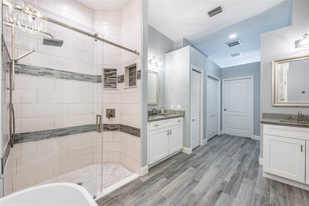 Primary en-suite luxurious rainfall shower with a frameless glass sliding barn door, dual niches, floor to ceiling tile surround and two separate vanities with sinks on Stylish wood-grained porcelain tile.