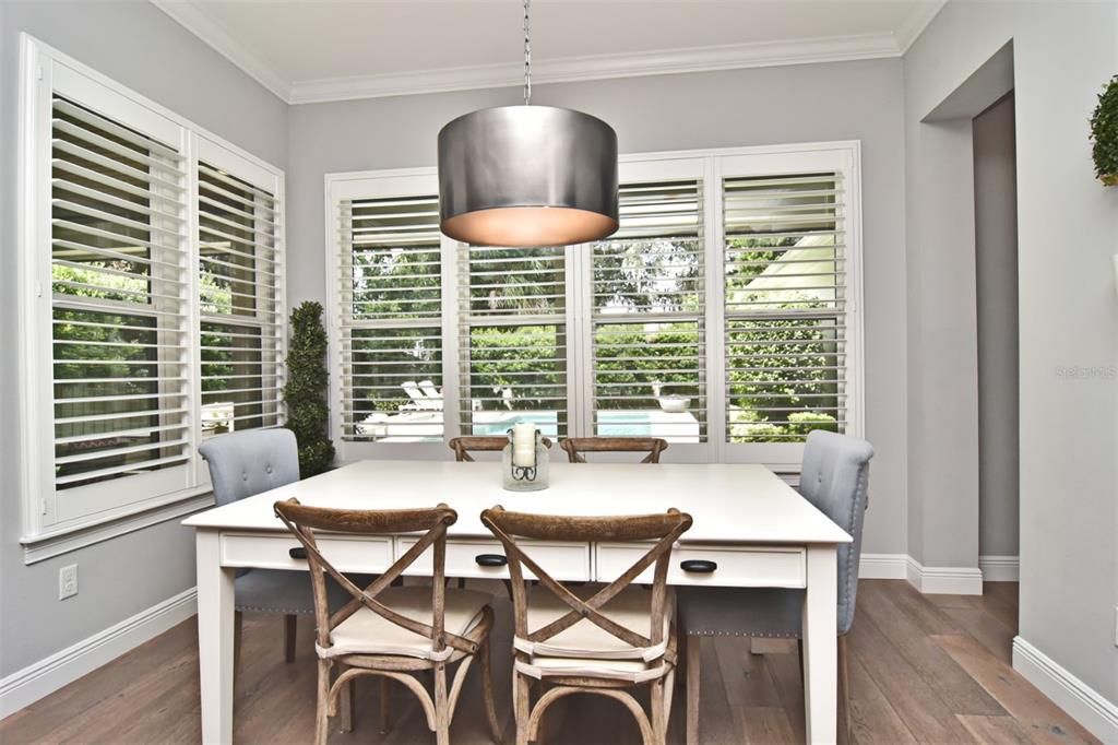 Dining area adjacent to kitchen, with wide-plank wood flooring, crown molding, and large windows overlooking pool and private backyard