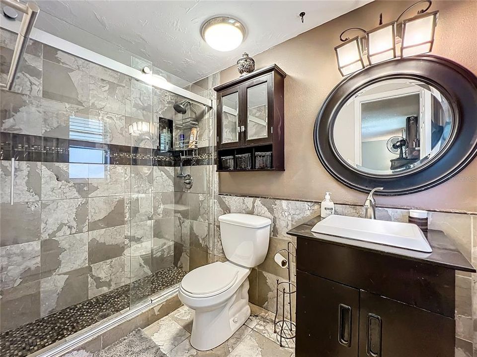 Primary bathroom with nicely updated shower, toilet, cabinets, mirror, light fixture, and floor tile.