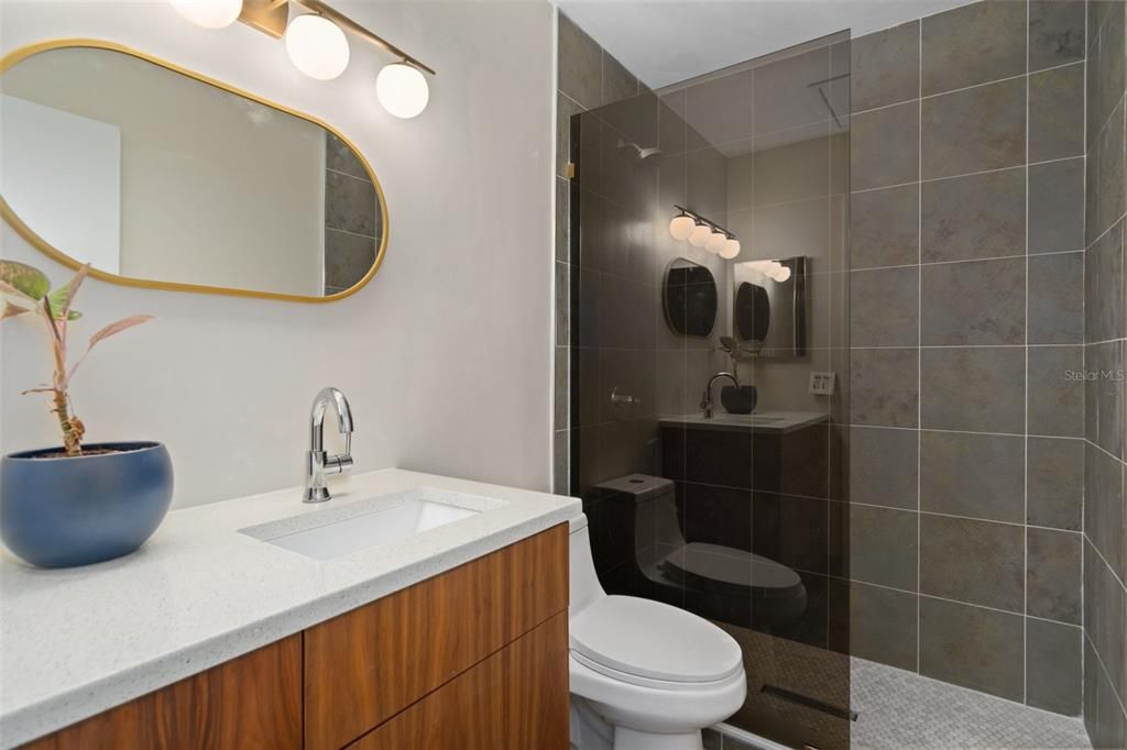 The bathroom features a floor to ceiling tiled, walk-in shower, a decorative mirror and light fixture above the vanity with storage and stone countertop.