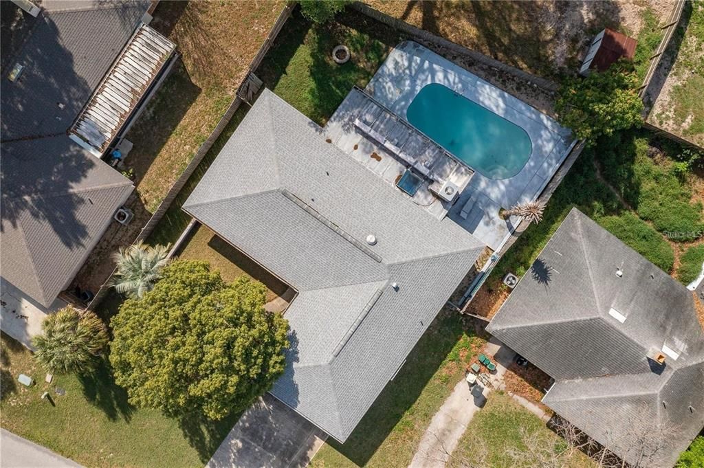 Drone photo showing the property from above, roof updated in 2021, 15x30 pool and Florida room, yard landscaping, wooden fence, driveway and street.