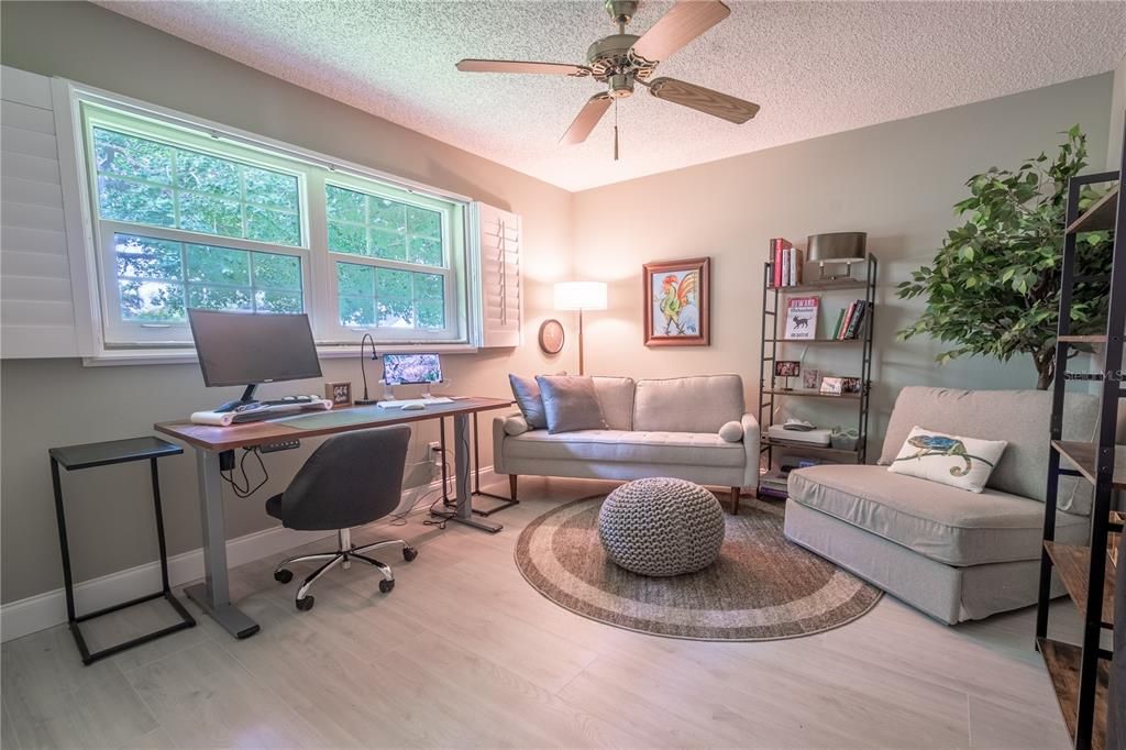 Bedroom 2 (currently used as a home office) is bathed in neutral tones, has wood laminate flooring, a ceiling fan with light kit and plantation shutters.