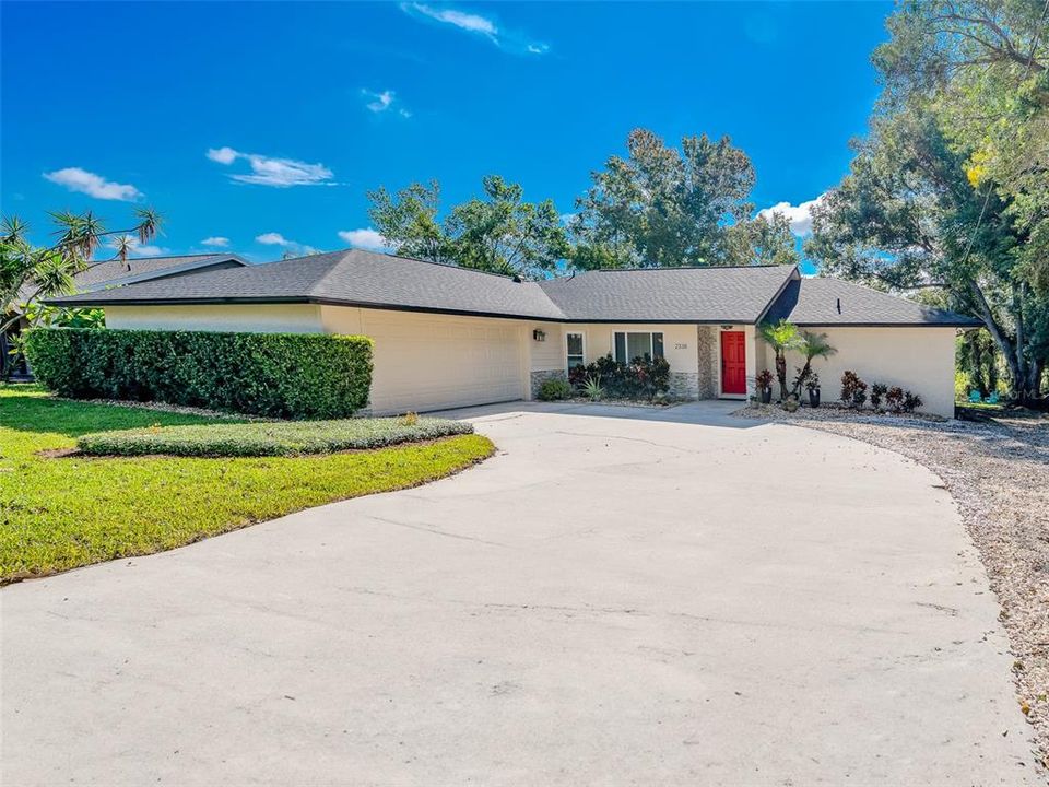 Spacious driveway leading to a well-maintained single-story home with a welcoming red front door. The lush greenery and mature landscaping offer privacy and curb appeal, making this home an inviting space to call your own.