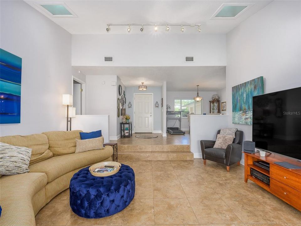 Spacious living area with high ceilings and natural light from skylights. The open floor plan connects to the entryway and dining area, creating an inviting and airy atmosphere. Tile flooring throughout adds durability and easy maintenance.