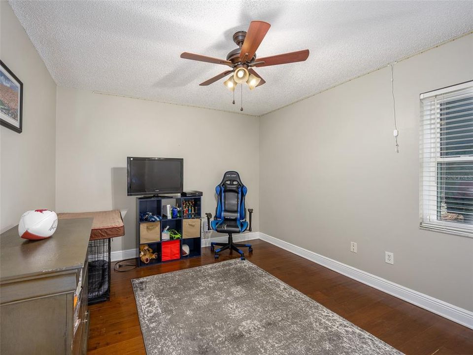 Spacious room with natural light and hardwood flooring, offering versatile use as a bedroom, playroom, or additional living space. Features a ceiling fan and large window, providing comfort and brightness. Ideal for customization to suit any need.