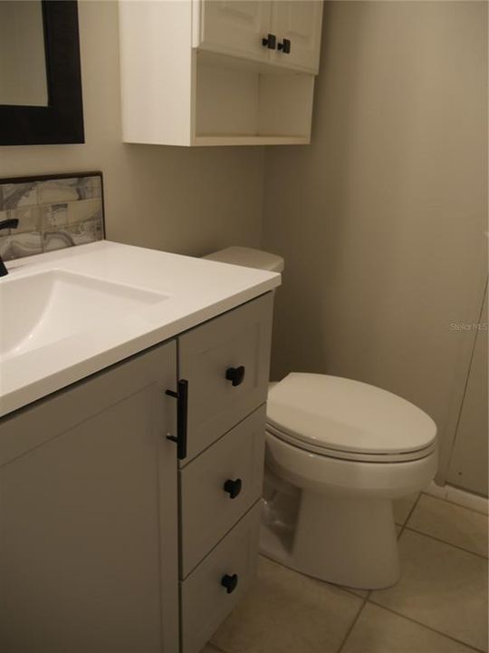 Primary bedroom's half-bath with new toilet, faucet, mirror, lighting, vanity and glass tile backsplash, with sink and solid surface counter.