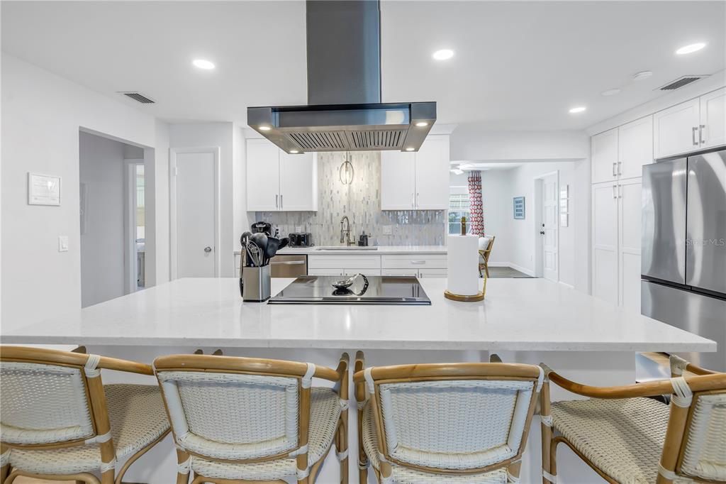 central island, rattan bar stools, a stainless steel range hood, and a sleek backsplash, blending into an open-concept dining and living area.