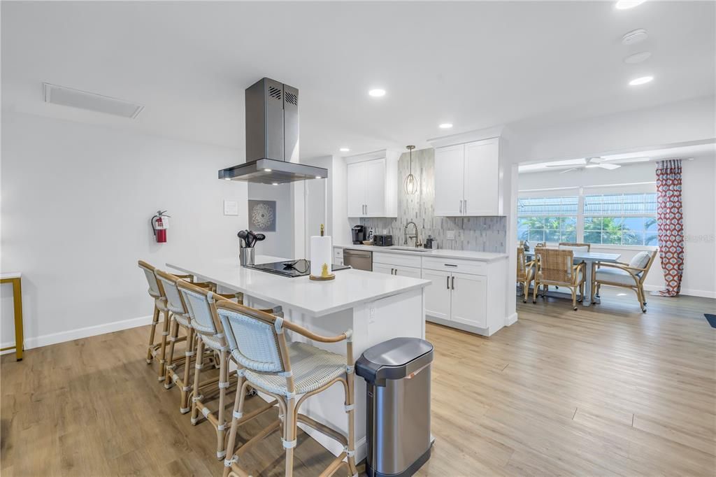 white island, stainless steel range hood, rattan bar stools, a mosaic tile backsplash, and a dining area in the background with large windows.