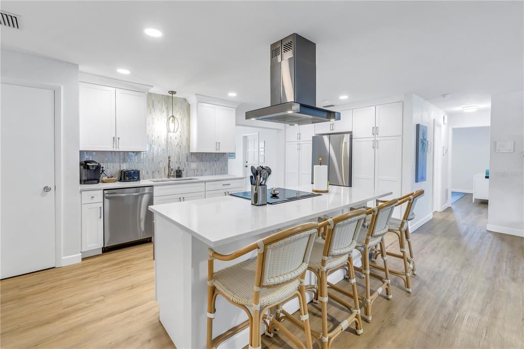 central island, rattan bar stools, a stainless steel range hood, and a sleek backsplash, blending into an open-concept dining and living area.