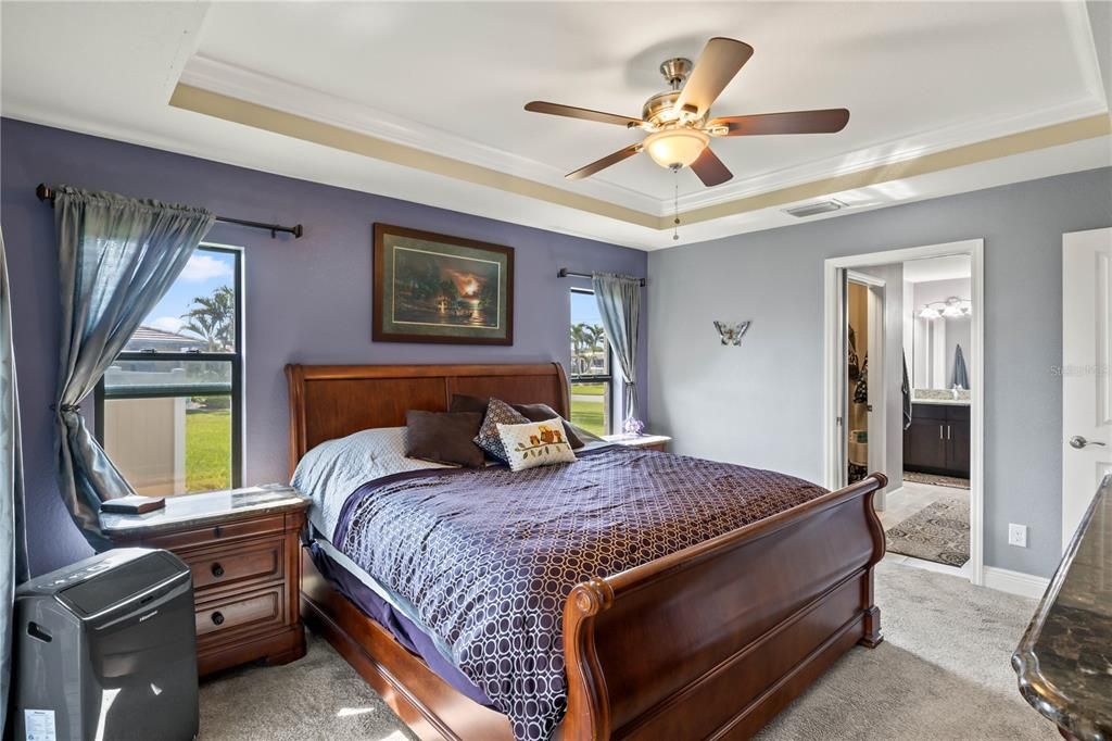 Carpeted bedroom with multiple windows, a tray ceiling, ensuite bath, and ceiling fan