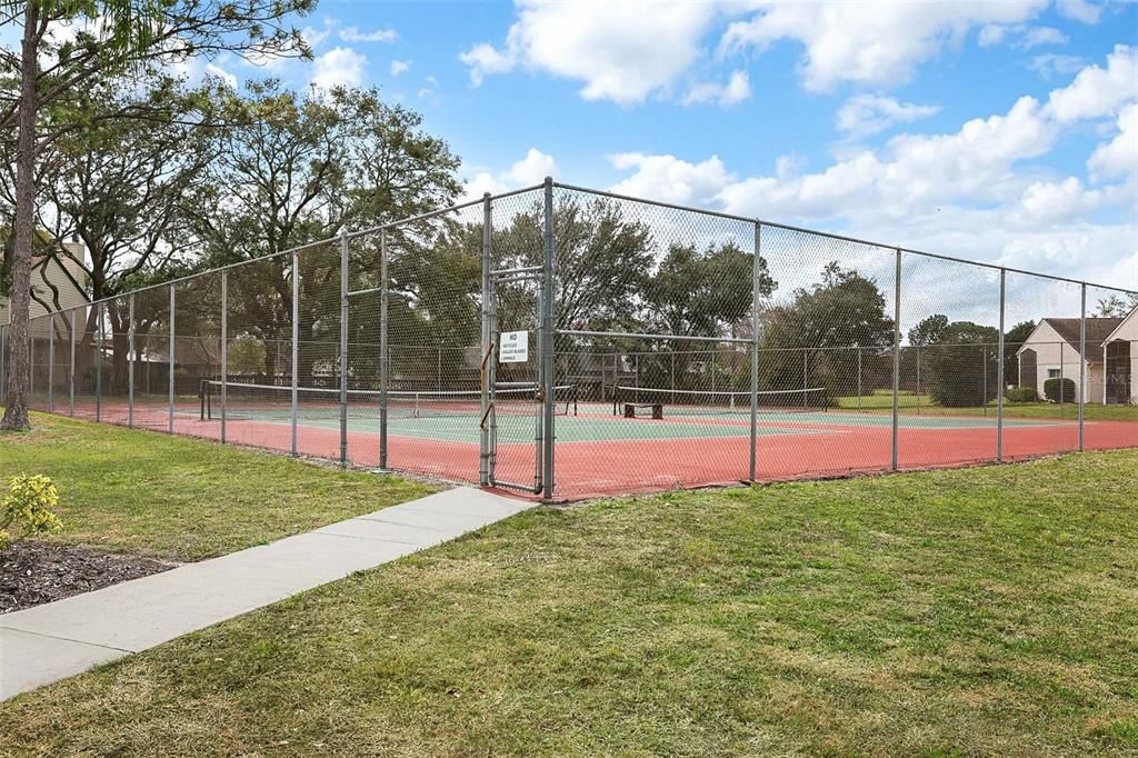 The image shows a fenced tennis court within a residential community. The court has a red surface with green playing areas and is surrounded by a chain-link fence. A sign on the gate reads "No Private Lessons Allowed." Inside, there are two tennis nets set up, providing space for multiple players. A concrete pathway leads up to the gate entrance, and the surrounding area features well-kept grassy lawns and landscaping. In the background, mature trees and residential buildings are visible, adding a peaceful, suburban feel to the recreational setting. The court offers residents a convenient and open space for outdoor activities.