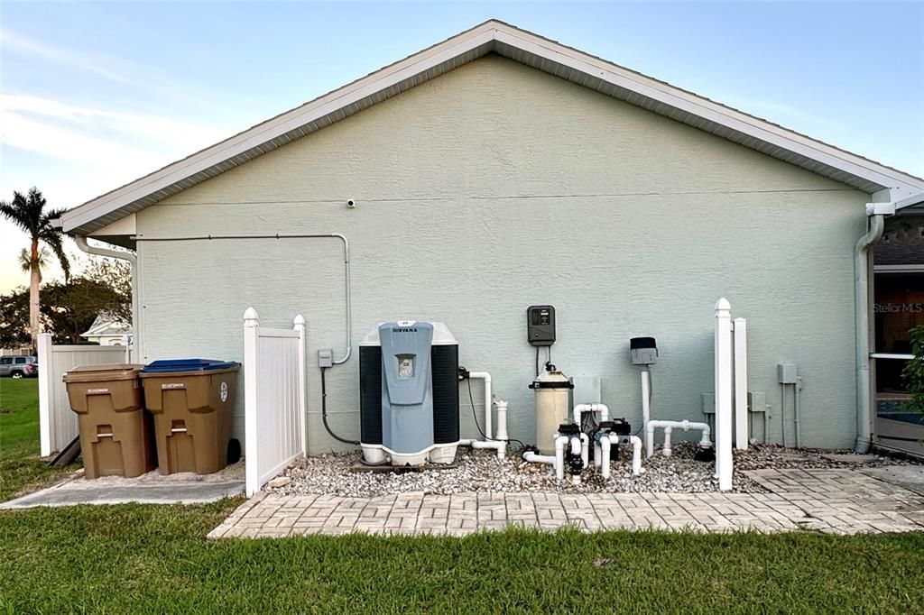 This back of the home features a comprehensive pool utility setup, including an electric heater, cartridge filter, saltwater panel and generator, as well as essential electric panels. An air blower enhances the spa experience, while clearly labeled valves allow for precise control of water flow throughout the pool system. A powerful pool pump maintains circulation, supported by floor cleaner valves to keep the pool pristine. Additionally, an in-line chlorine feeder is available, though it is not used within the saltwater system. A privacy screen partially encloses the equipment, keeping the area tidy and visually unobtrusive. Additionally, the setup includes a convenient space for waste and recycling bins. The entire arrangement is set against the clean exterior of the home, with gravel for easy maintenance and a paved walkway for access.