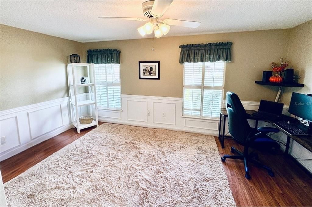 This bright and spacious second bedroom offers a comfortable and versatile layout, perfect for creating a personalized retreat. Elegant wainscoting along the walls adds a touch of sophistication, complementing the warm wood-style flooring. Large windows with plantation shutters provide ample natural light and offer a view of the serene outdoors, while the overhead ceiling fan with a light fixture ensures comfort year-round.The room’s neutral color palette and large walk-in closet make it easy to adapt to various decor styles, whether for a bedroom, home office, or creative space. With direct access to other areas of the home, this room combines both privacy and convenience. The blend of classic details and functional design creates a welcoming and adaptable space suited to many uses.